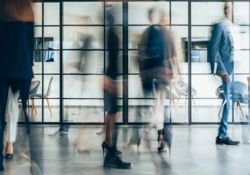 Office corridor with glass partitions and blurred people walking, showing movement and an active workplace atmosphere.
