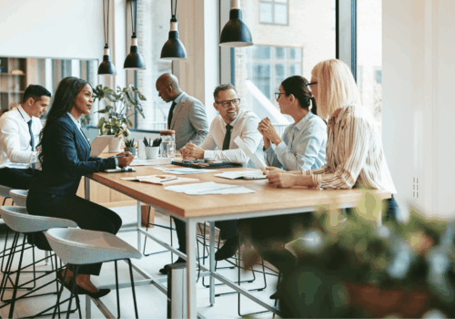Team meeting around a high table in a bright office, discussing documents in a collaborative workspace.