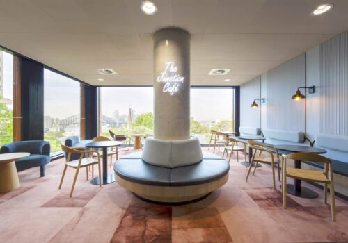 Office cafe with circular seating around a column, timber chairs, soft furnishings, and city views through windows.