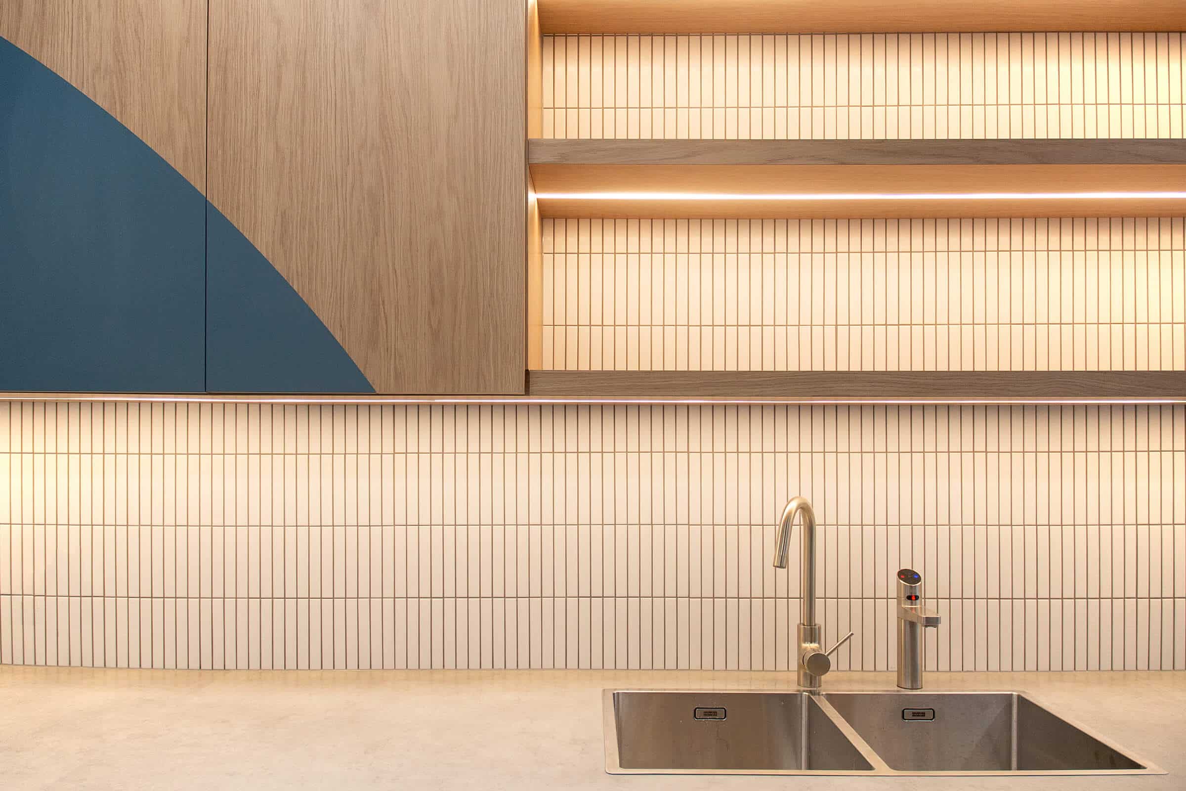 Kitchen splashback detail with vertical tiled pattern, timber shelving, integrated lighting, and stainless steel sink.