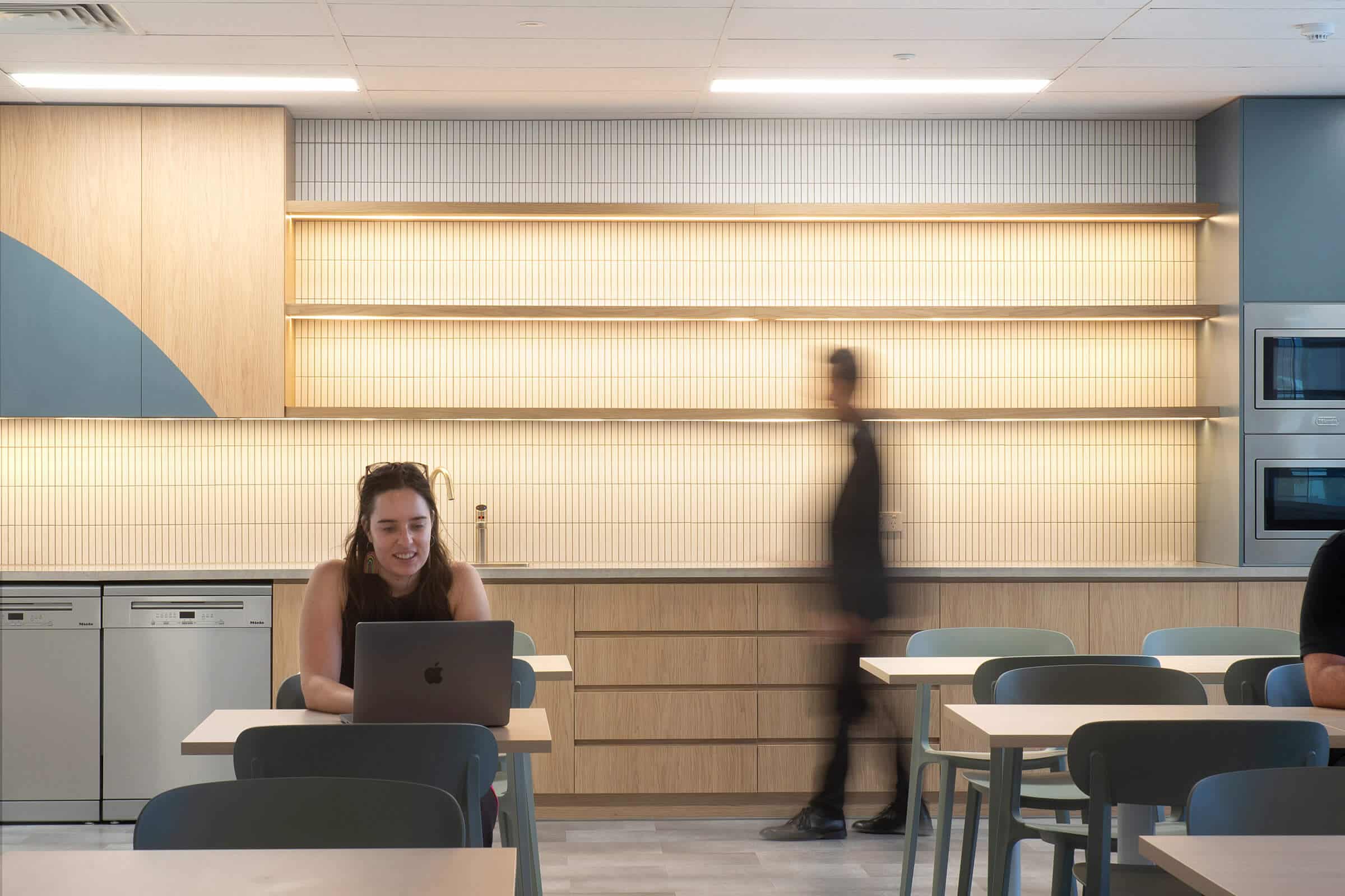 Staff breakout space with tables and chairs, timber cabinetry, and softly lit shelving, with people moving through.