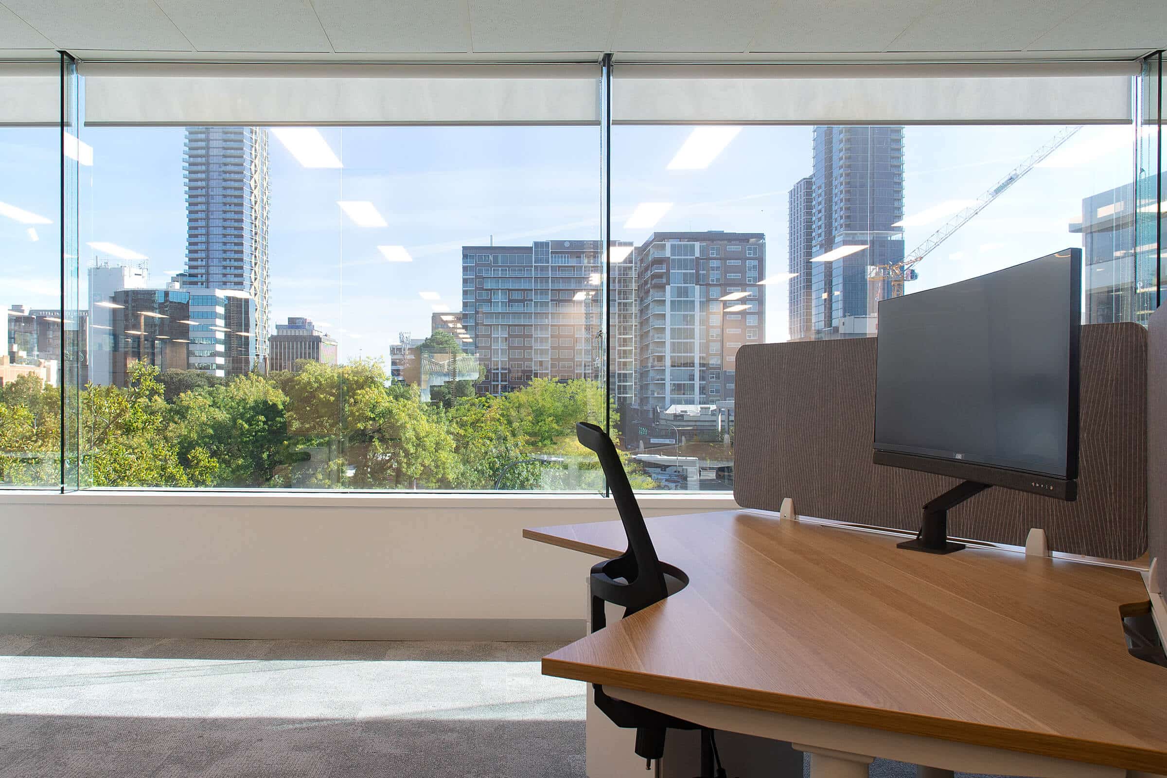 Workstation desk with monitor positioned beside full height windows overlooking trees and surrounding city buildings.