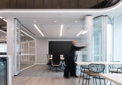 Meeting room with sliding glass walls, conference table, ceiling lighting and adjacent breakout seating.